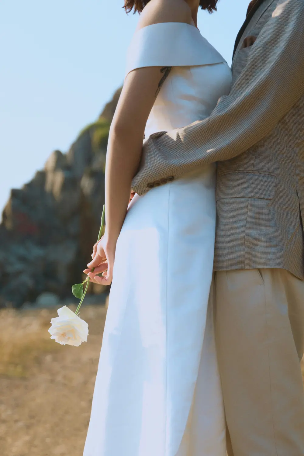 Mariée en robe blanche tenant une rose blanche dans le dos, enlacée par le marié en veste beige, devant une falaise sous un ciel bleu