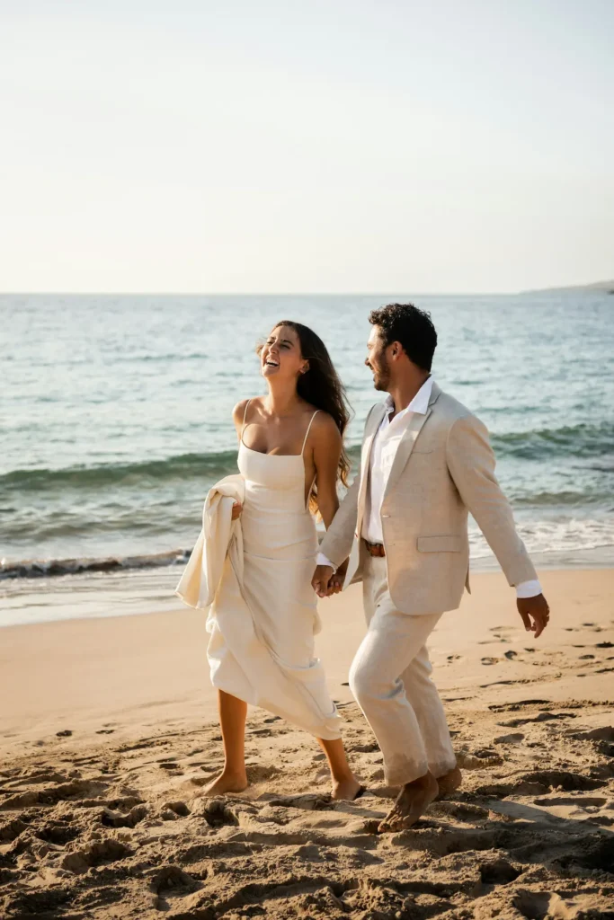 Mariés main dans la main, pieds nus sur le sable au bord de la mer, la mariée en robe crème riant aux éclats à côté du marié en costume de lin beige