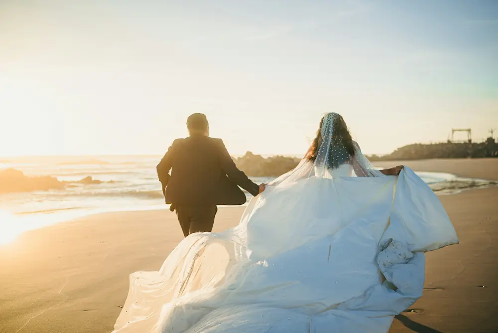Couple de mariés de dos courant main dans la main sur la plage au coucher du soleil, long voile orné de perles flottant derrière la mariée