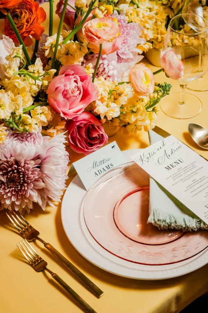 Table de mariage dressée sur une nappe jaune avec centre de table de renoncules, roses et chrysanthèmes roses et orangés, assiette en verre rose, serviette vert sauge, menu et marque-place calligraphiés